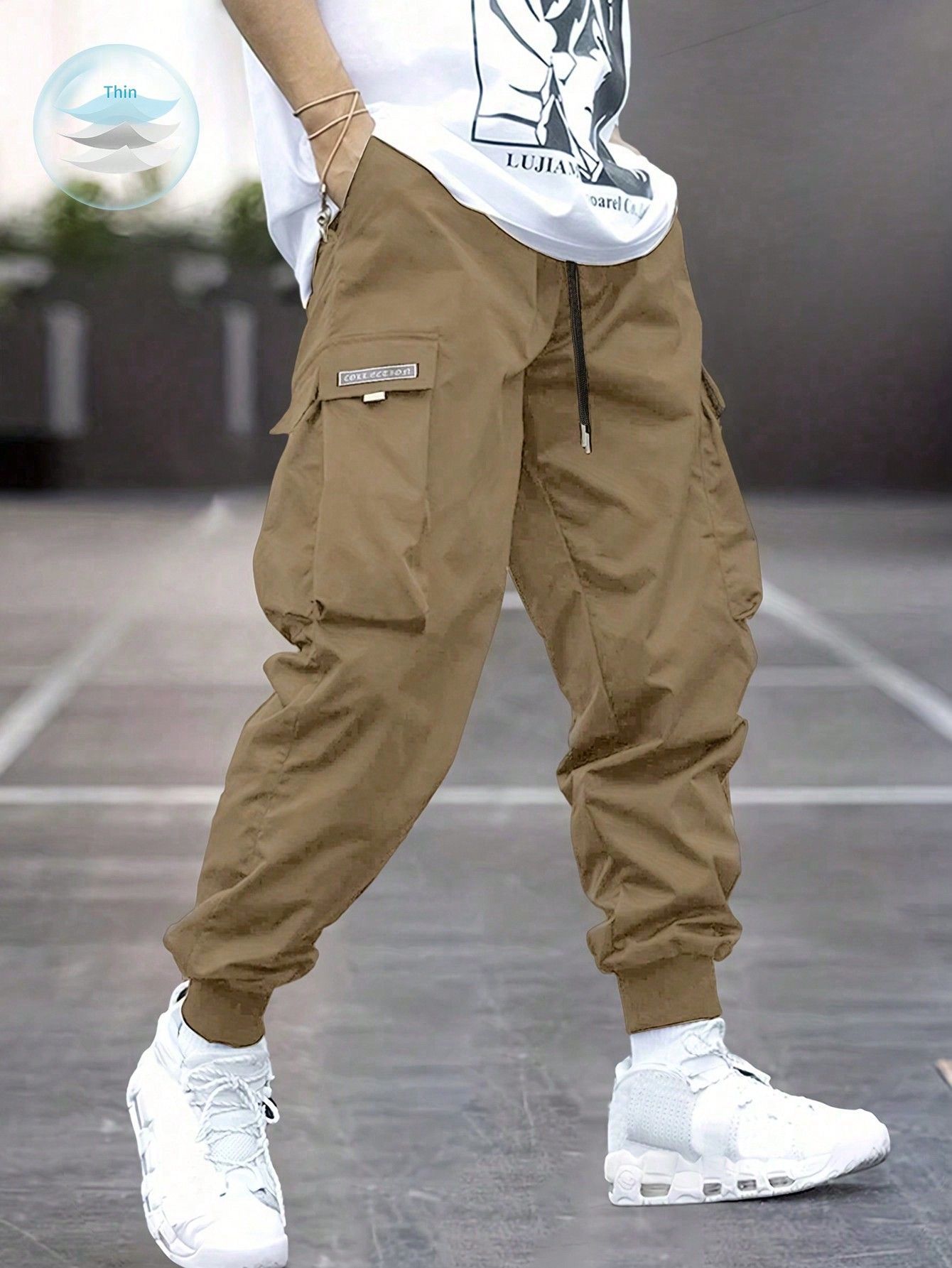 A man wearing slim-fit khaki cargo pants in a warm tan/beige/khaki color. The pants feature a tapered, jogger-style leg with elastic cuffs at the ankles, multiple large flap cargo pockets on the thighs (with visible stitching drawstring details at the waist, and a relaxed yet fitted silhouette through the hips and thighs. He’s paired them with white sneakers and a white graphic t-shirt that’s slightly cropped or tucked, standing on a tiled indoor floor in a casual streetwear pose from mid-thigh down