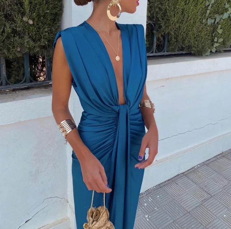 Woman wearing a blue dress with gold accessories against a white wall.